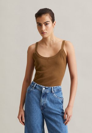 Frau mit zurückgebundenem dunklem Haar, die ein braunes, geripptes Tanktop und hochtaillierte blaue Jeans vor einem schlichten hellen Hintergrund trägt.