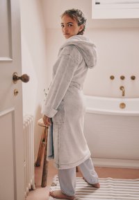 Accappatoio grigio in morbido tessuto con cappuccio, tasche laterali, indossato sopra pantaloni del pigiama a righe blu e bianche. Texture soffice, design semplice e vestibilità casual.