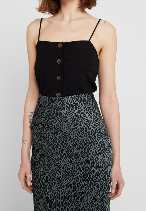 Femme portant un haut noir à bretelles fines boutonné et une jupe taille haute à imprimé léopard vert, avec des tatouages sur son bras gauche.