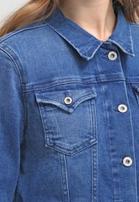 Tmavě modrá džínová bunda s klasickým límcem, předními kapsami, stříbrnými patentkami a viditelnými švy. Texturovaný povrch látky.