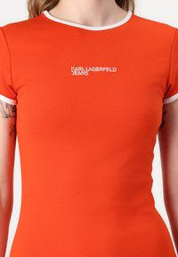 Rdeča prilega majica s kratkimi rokavi z belim rebrastim ovratnikom in rokavi. Ima vezeno oznako "KARL LAGERFELD JEANS" spredaj.