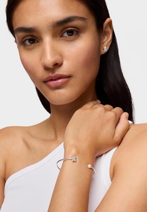 Młoda kobieta o ciemnych włosach, nosząca srebrne kolczyki w kształcie niedźwiedzi oraz pasującą, otwartą srebrną bransoletkę z perłowym akcentem, ubrana w biały, jedno-ramienny top.
