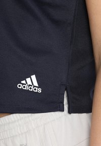 Canotta sportiva blu navy con logo "adidas" bianco. Caratterizzata da un tessuto texture e fessure laterali per la mobilità. Indossata con pantaloni corti chiari.