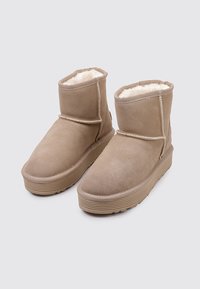 Par de botas de tobillo de ante beige con suelas de goma gruesas y forro de lana blanca, presentadas sobre un fondo gris claro.
