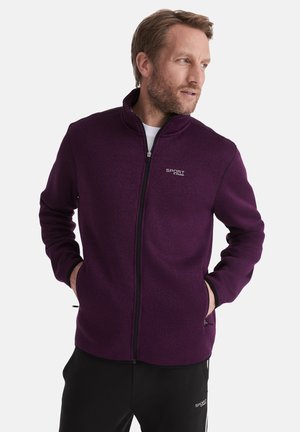 Mann in einer lila Zip-Jacke mit "SPORT"-Logo, mit Händen in den Taschen, schaut zur Seite, vor einem schlichten weißen Hintergrund.