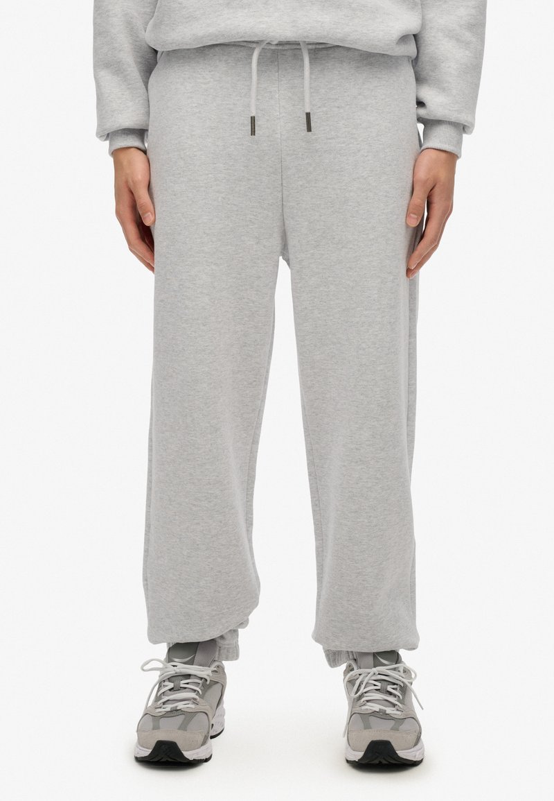 Pantalon de survêtement gris clair avec poignets élastiques et taille à cordon, porté avec des chaussures de sport grises et blanches sur fond blanc.