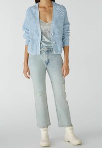 Vaaleansininen neulottu neuletakki, jossa on resoritekstuuri, napit edessä, puettuna hopeisen paljettitopin päälle ja yhdistettynä vaaleansinisiin farkkuihin sekä valkoisiin saappaisiin.