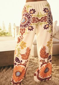 Pantaloni larghi di colore crema con grandi motivi floreali in arancione, blu, verde e bordeaux, dotati di una vita elastica.