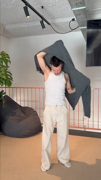 Personne portant un pantalon blanc et un débardeur enfilant un pull gris à l'intérieur près d'un pouf et d'une plante.