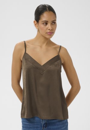 Femme aux cheveux foncés coiffés en un chignon bas, portant un caraco en satin marron à fines bretelles et un jean bleu, debout devant un fond uni.