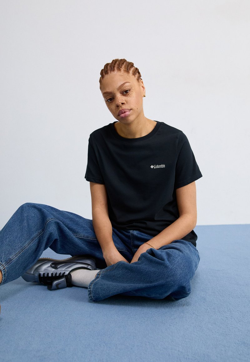 Schwarzes Baumwoll-T-Shirt mit kurzen Ärmeln und kleinem Columbia-Logo. Große blaue Jeans, graue Sneaker und hellgraue Socken. Sitzt auf blauem Boden.