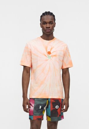 Młody mężczyzna z warkoczami, ubrany w jasnopomarańczową koszulkę tie-dye i wielokolorowe krótkie spodenki z grafiką, stojący na jednolitym białym tle.