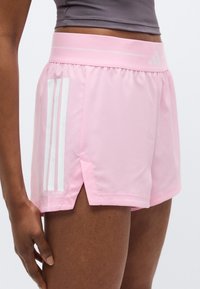 Ροζ αθλητικό σορτς με ελαφρύ ύφασμα, με λευκό σχεδιασμό τριών γραμμών, πλευρικές σχισμές και ελαστική μέση.