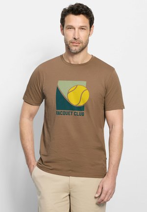 Man met baard draagt bruin "Racquet Club" t-shirt met tennisbal afbeelding, beige broek, staat met één hand in de zak.