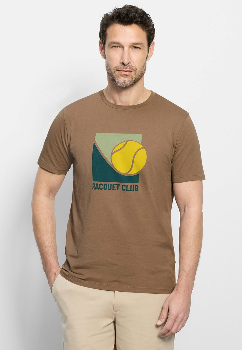 Homme avec barbe portant un t-shirt marron "Racquet Club" avec un motif balle de tennis, pantalon beige, debout avec une main dans la poche.