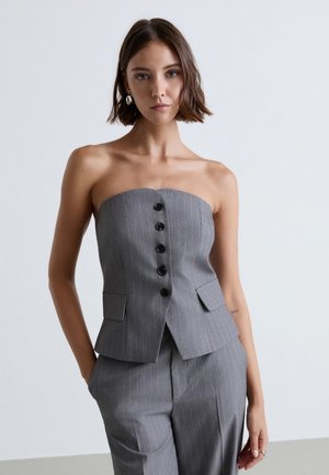 Donna che indossa un top grigio a righe sottili senza spalline con bottoni neri e pantaloni a vita alta abbinati, in piedi davanti a uno sfondo chiaro e semplice.