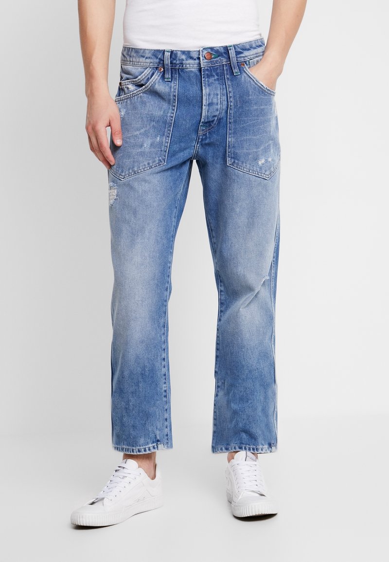 Mies, joka on pukeutunut vaaleansinisiin kuluneisiin farkkuihin, joissa on suuret etutaskut, ja valkoisiin lenkkareihin, seisoo yksinkertaisen taustan edessä.