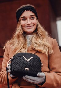 Schwarze gesteppte Handtasche mit roségoldenen Akzenten, runder Form und verstellbarem Schultergurt, gehalten von einer Person mit grauen Handschuhen vor einem verschwommenen Hintergrund.