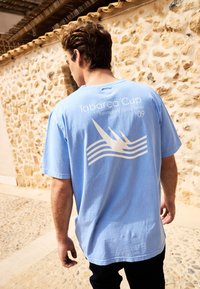 Ljusblå bomull T-shirt med korta ärmar, med vit text och en grafisk design av vågor och segel på baksidan.