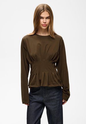 Kvinde med glat, skulderlangt hår iført en langærmet, tætsiddende olivengrøn top og mørkeblå jeans mod en ensfarvet baggrund.