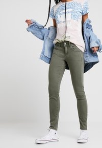 Olivově zelené džíny s úzkým střihem, kombinované se světle modrou denimovou bundou a pastelovým květinovým tričkem. Bílé plátěné tenisky dokončují vzhled.