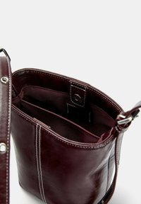 Temno rjav usnjen crossbody nahrbtnik z belimi šivi, nastavljivim trakom in notranjo tkaninsko oblogo z več predali ter zaklepanjem na gumb.
