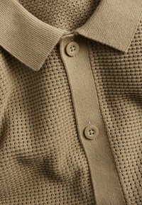 Beige gebreide poloshirt close-up dat de textuur van de stof, de geribbelde kraag, de sluiting en twee ronde knopen toont.