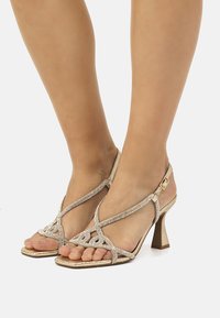 Sandalias de tacón color dorado con acentos de cristal, diseño con tiras, estilo de punta abierta y un tacón moderado de bloque. Textura suave y forma elegante.
