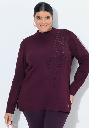 Kvinde iført en mørkelilla langærmet sweater med dekorative perlemønstre, kombineret med matchende lilla bukser, smilende til kameraet.