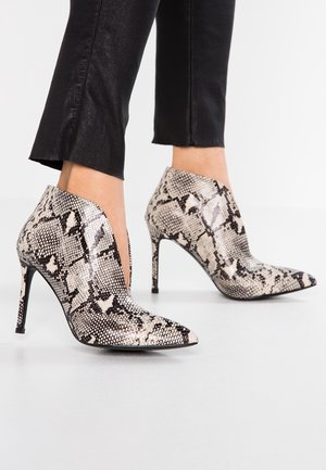 Femme portant des bottines à bout pointu avec un motif peau de serpent noir et blanc et des talons fins et hauts, associées à un pantalon noir court.