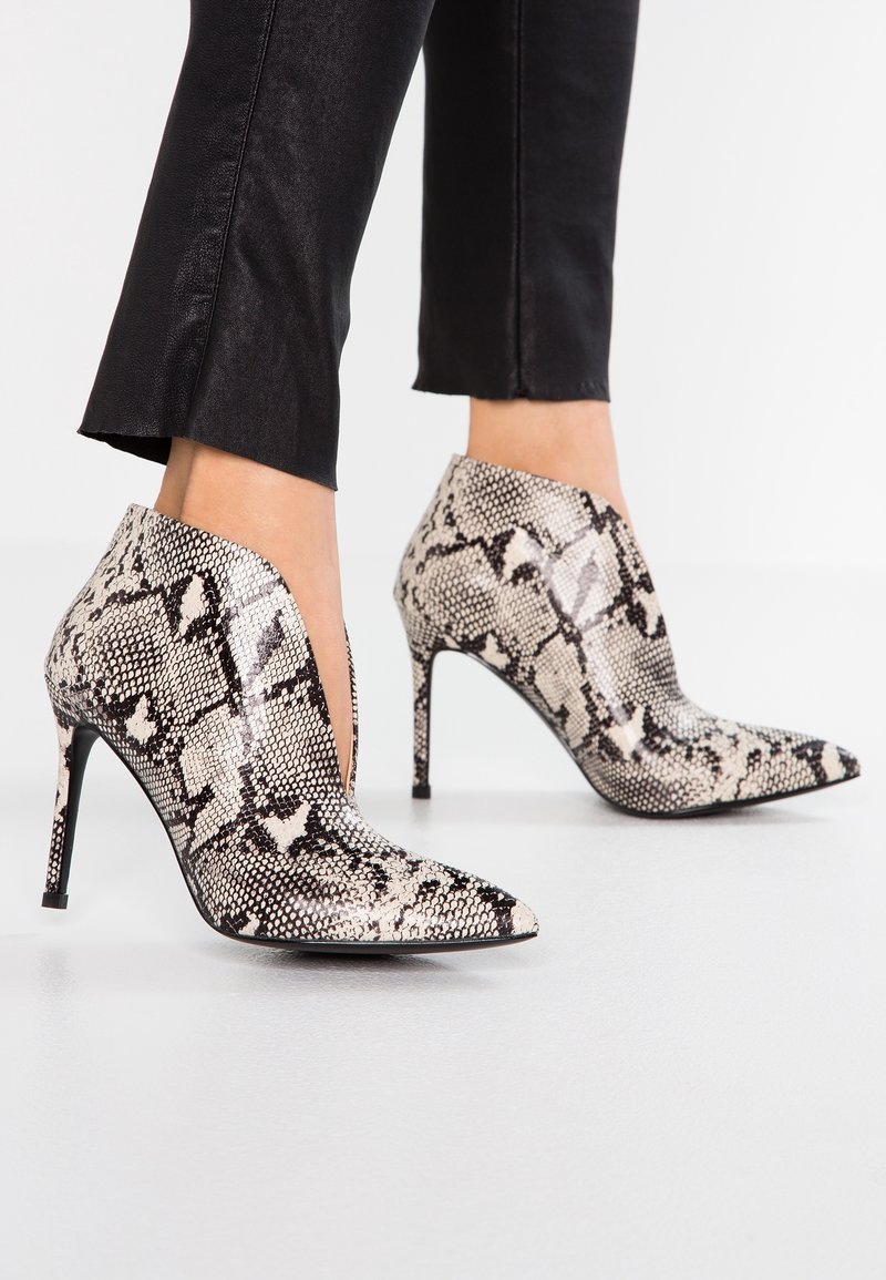 Femme portant des bottines à bout pointu avec un motif peau de serpent noir et blanc et des talons fins et hauts, associées à un pantalon noir court.