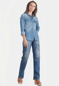 Chemise en denim bleu clair avec boutonnage sur le devant et deux poches poitrine ; associée à un jean bleu foncé avec délavage et fentes latérales.