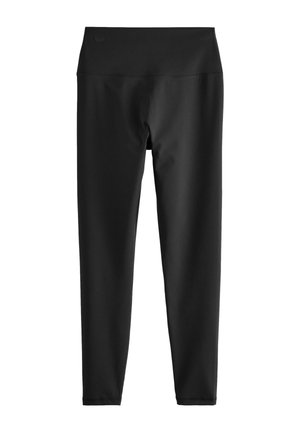 Vrouw draagt zwarte legging met hoge taille en het logo "self." op de tailleband en een groen-wit gestreept shirt, handen rustend op heupen en dij.