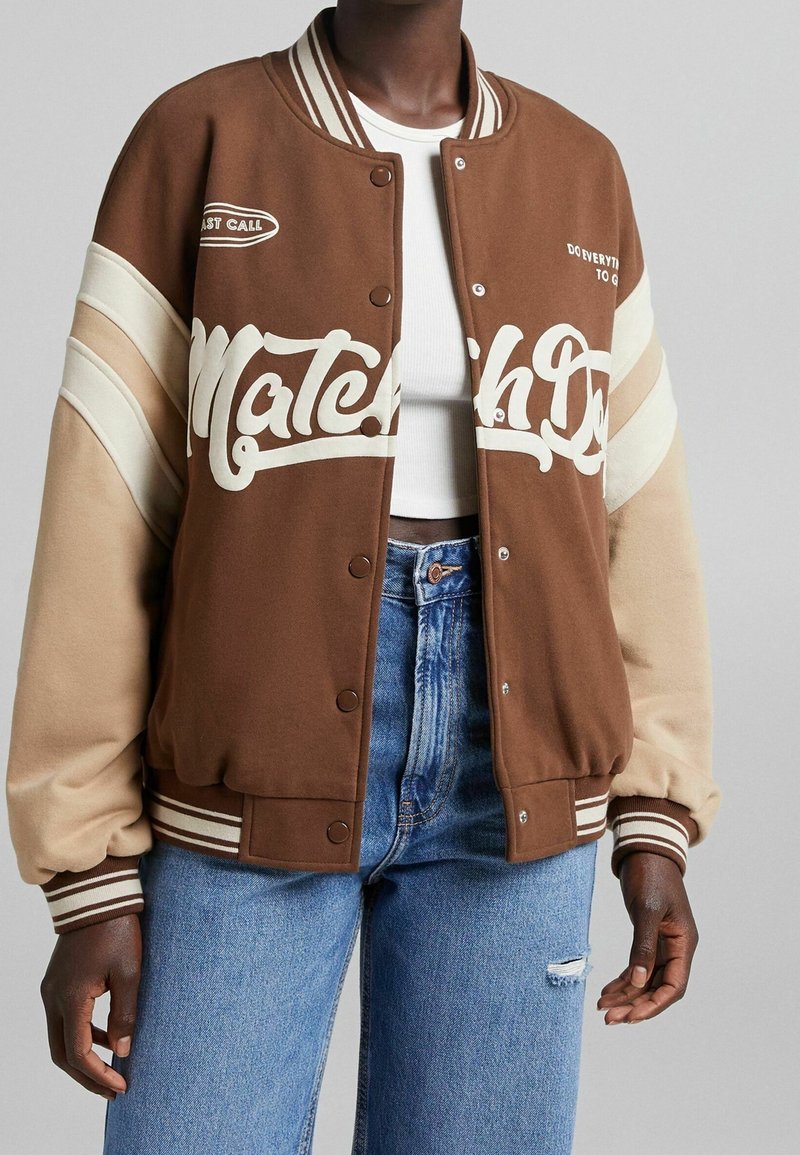 Brun varsityjacka med vit text, beige ränder, ribbade muddar och tryckknappar. Bärs över en vit topp och kombineras med blå jeans.