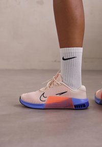 Zapatillas deportivas Nike en malla rosa claro con un degradado de acentos naranjas y azules. Presentan el logo Swoosh negro y calcetines blancos de acanalado.
