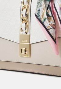 Sac à main blanc à motif géométrique avec une chaîne dorée et une écharpe en soie colorée. Doté d'un fermoir doré et d'une base texturée beige.