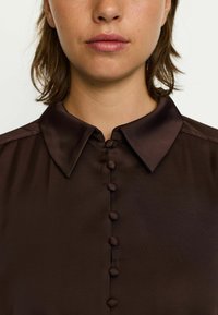 Hnedá blúzka s klasickým golierom, texturovanou tkaninou a predným zapínaním na pät gombíkov, s hladkým povrchom.