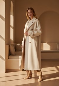 Cremefarbener Wollmantel mit doppelter Knopfleiste, großen Knöpfen und taillierter Silhouette. Kombiniert mit beigen High-Heels-Stiefeln.