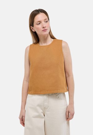 Femme aux cheveux bruns lisses portant un haut sans manches couleur camel et un pantalon beige taille haute, regardant légèrement vers sa gauche.