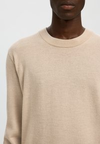 Nærbillede af en person iført en beige crewneck-trøje, der viser skuldre, bryst og en del af nakken og den nedre del af ansigtet.