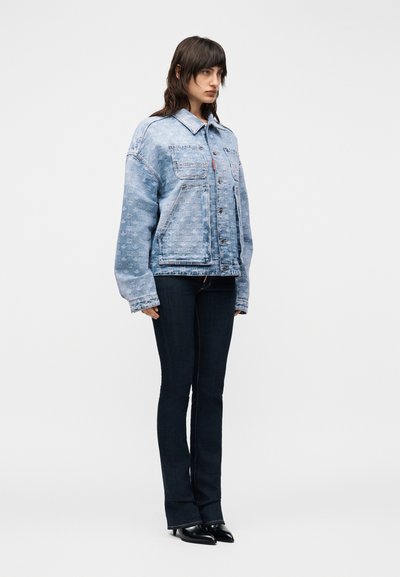 Femme portant une veste en denim bleu clair à motifs oversized, un jean foncé ajusté et des chaussures noires à bouts pointus, debout devant un fond uni.