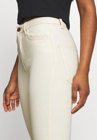 Zblízka na osobu v krémových džínách s vysokým pasem, s viditelnými předními a zadními kapsami, hnědým knoflíkem a černým zkráceným topem.