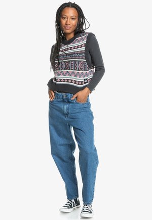 Gebreide sweatervest met geometrische patronen in roze en grijs, gecombineerd met hoge blauwe spijkerbroek en zwarte canvas sneakers.