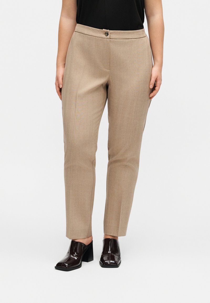 Μπεζ παντελόνι σε μήκος μέχρι τον αστράγαλο, σχεδιασμένο κατά παραγγελία, φορεμένο με μαύρο τοπ και σκούρα καφέ δερμάτινα παπούτσια με τακούνι, στέκεται σε απλό λευκό φόντο.