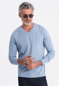 Hellblaues langärmliges V-Ausschnitt-Shirt aus Baumwollmischung mit gerippten Ärmelabschlüssen, kombiniert mit dunkelblauen Jeans und dunklen, runden Sonnenbrillen.