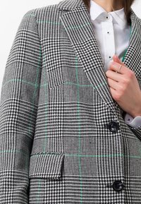 Blazer a scacchi realizzato in tessuto testurizzato bianco e nero con dettagli verdi, caratterizzato da un colletto a rever e chiusura a due bottoni.