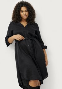 Schwarzes Leinenkleid mit durchgehender Knopfleiste, Kragen und einem Gürtel in der Taille; hat Dreiviertelärmel und einen etwas längeren hinteren Saum.