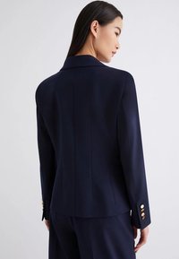 Blazer navy con una silhouette aderente, colletto affilato e dettagli con bottoni dorati sulle maniche. Il tessuto liscio e i dettagli delle cuciture verticali esaltano il design.