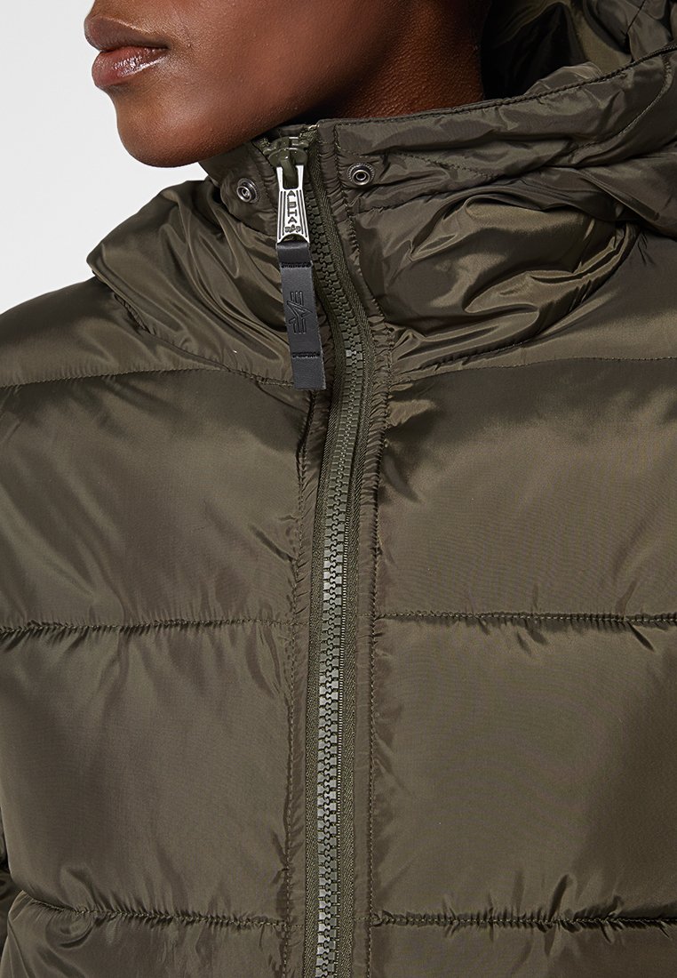 Primer plano de una persona que lleva una chaqueta de plumas verde olivo con capucha y un tirador de cremallera de marca.