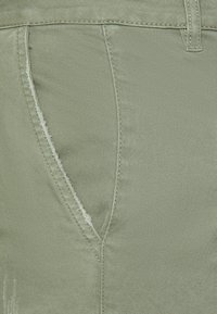 Tecido de ganga verde com uma textura suave, apresentando um bolso frontal e costura visível. Inclui um detalhe desgastado perto do bolso.